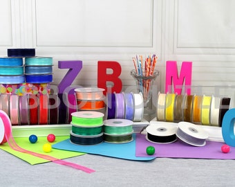 May include: Assortment of colourful ribbon spools in various colours and textures, including pink, blue, green, yellow, orange, and white. The spools are arranged on a white surface with colourful letters and a vase of straws.