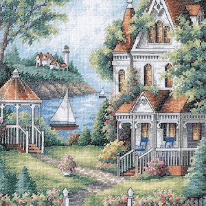 May include: A cross-stitch landscape depicts a coastal scene with a white house, gazebo, and sailboat on the water. The house has a brown roof and is surrounded by lush greenery and flowers. A lighthouse is visible in the distance.