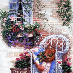 May include: A cross-stitch depiction of a serene outdoor scene. A white wicker chair with a patterned blanket and a basket of roses sits near a brick wall with a window box overflowing with flowers. Potted plants add color.