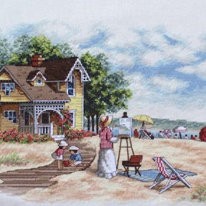 May include: Cross-stitch depicting a beach scene with a yellow house, people, and a woman painting. The house has a brown roof and white trim. A woman in a striped shirt paints on an easel. Two children play nearby. Beach umbrellas and a red and white striped chair are visible.