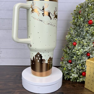 Op de afbeelding: Een witte en gouden roestvrijstalen reistumbler met een handvat en een deksel. De tumbler heeft een wintersc&egrave;ne met rendieren die een slee trekken en een skyline van een stad. De tumbler staat op een witte draaitafel.