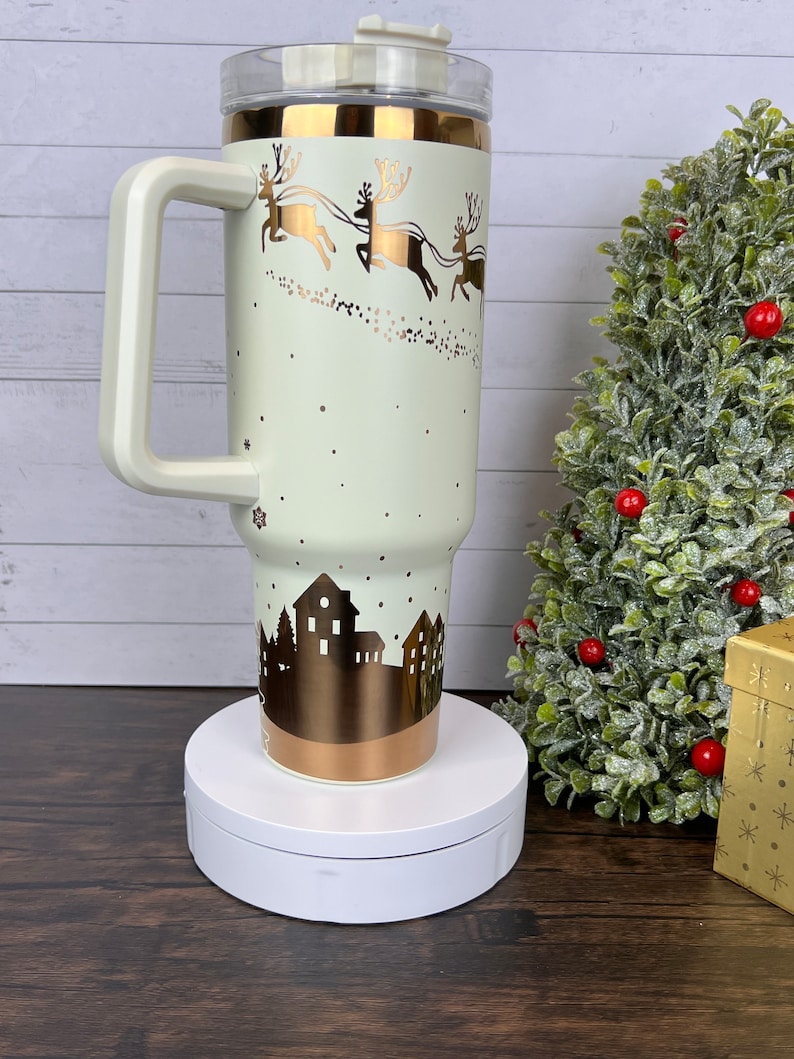 Op de afbeelding: Een witte en gouden roestvrijstalen reistumbler met een handvat en een deksel. De tumbler heeft een wintersc&egrave;ne met rendieren die een slee trekken en een skyline van een stad. De tumbler staat op een witte draaitafel.