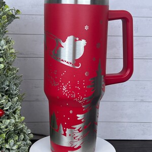 Op de afbeelding: Rode roestvrijstalen reistumbler met een handvat. De tumbler heeft een lasergegraveerd ontwerp van Sinterklaas in een slee die over een besneeuwde bos sc&egrave;ne vliegt.