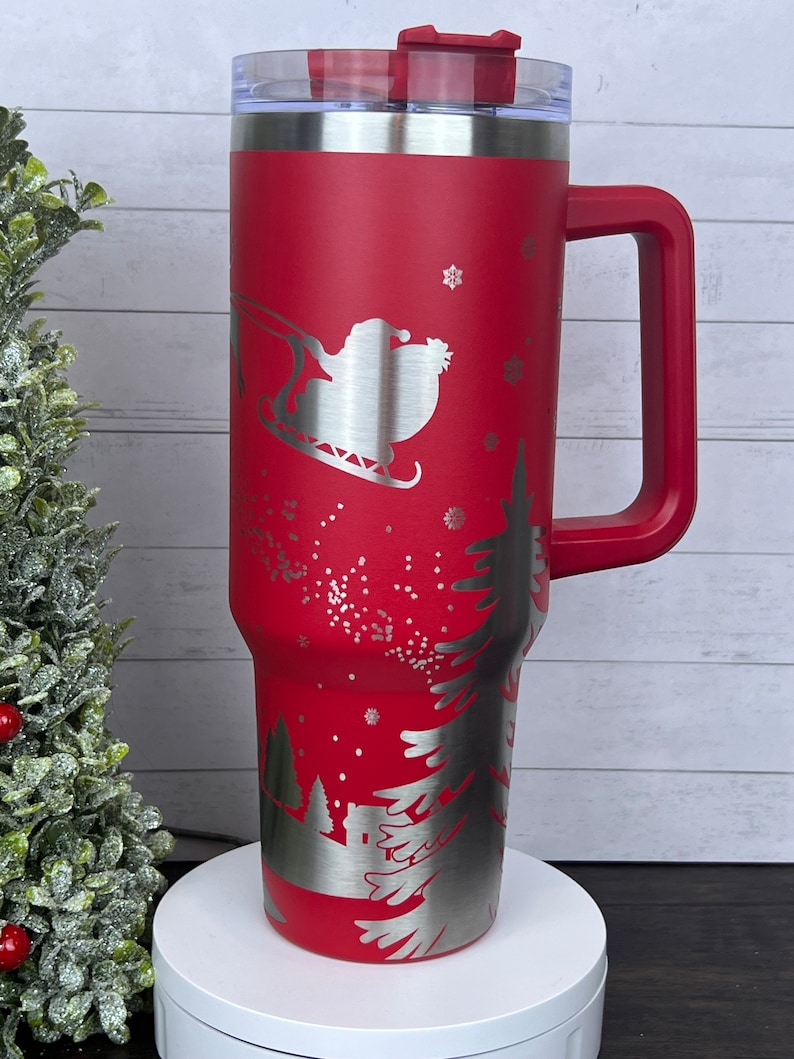 Op de afbeelding: Rode roestvrijstalen reistumbler met een handvat. De tumbler heeft een lasergegraveerd ontwerp van Sinterklaas in een slee die over een besneeuwde bos sc&egrave;ne vliegt.