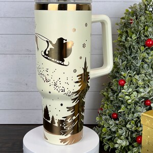 Op de afbeelding: Een witte reistumbler met een gouden handvat en een gouden kerstsc&egrave;neontwerp. Het ontwerp bevat Sinterklaas in een slee getrokken door rendieren, sneeuwvlokken en dennenbomen.