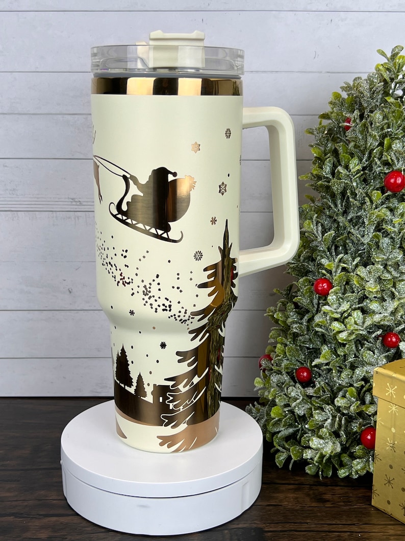 Op de afbeelding: Een witte reistumbler met een gouden handvat en een gouden kerstsc&egrave;neontwerp. Het ontwerp bevat Sinterklaas in een slee getrokken door rendieren, sneeuwvlokken en dennenbomen.