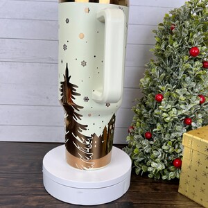 Op de afbeelding: Een witte en gouden roestvrijstalen beker met een wintersc&egrave;neontwerp. De beker heeft een deksel en een handvat. Hij staat op een witte draaitafel.