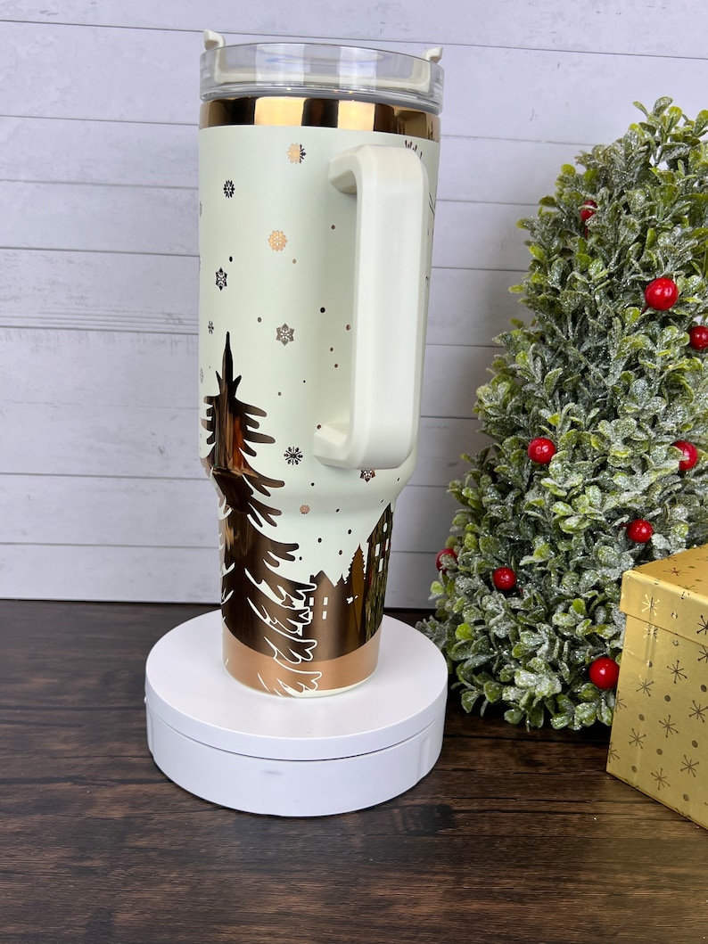 Op de afbeelding: Een witte en gouden roestvrijstalen beker met een wintersc&egrave;neontwerp. De beker heeft een deksel en een handvat. Hij staat op een witte draaitafel.