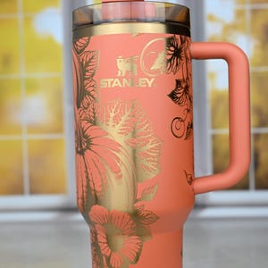 May include: A coral-colored stainless steel travel mug with a gold-colored pumpkin and leaf design. The mug has a handle and a lid with a straw.