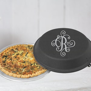 May include: A black pie plate with a silver monogram of the letter "R" on the lid. The pie plate is filled with a quiche.