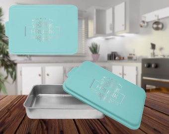 May include: A rectangular baking pan with a light blue lid. The lid has the text "MADE WITH LOVE" and "HRUBIK" in silver lettering. The pan is made of stainless steel and sits on a wooden surface.