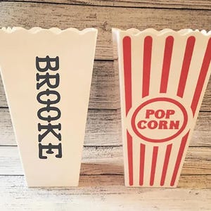 May include: Two white cardboard popcorn containers. One container has red and white stripes and says "POP CORN" in a circle. The other container has the word "BROOKE" printed in black.