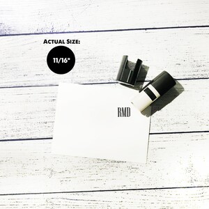 以下が含まれることがあります： 白地に「RMD」のイニシャルが刻まれた黒い自動インクスタンプ。スタンプの直径は1 1/16インチ。スタンプは木目調の背景の白いカードの上に置かれています。