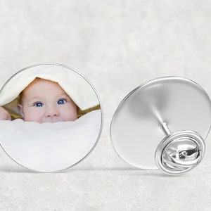 May include: A silver pin with a round image of a baby peeking out from under a white blanket. The pin has a safety clasp on the back.