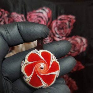 May include: A red, white, and black glass pendant with a swirling design. The pendant is round and has a hole at the top for hanging.