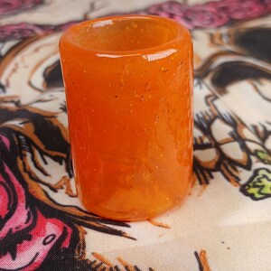 May include: A cylindrical orange glass object with glitter particles. The object is a short, open-ended tube, likely a shot glass or small container. The glass has a translucent quality, allowing light to pass through. The background is a patterned fabric.
