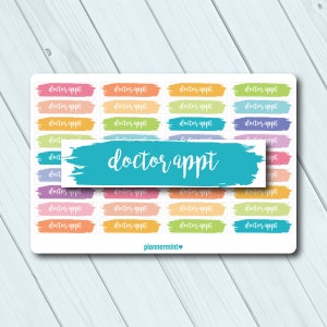 May include: A sheet of colorful planner stickers with the text "doctor appt" in white. The stickers are on a white background with a light blue wooden texture. The stickers are in various colors, including pink, orange, yellow, green, blue, and purple.