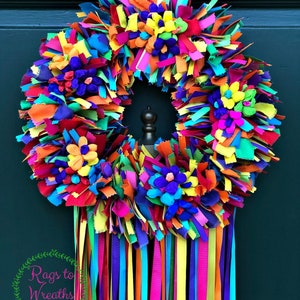 May include: A colorful rag wreath with long streamers in shades of pink, blue, green, yellow, and orange. The wreath is decorated with paper flowers in various colors. The wreath is hanging on a dark blue door. The text "Rags to Wreaths Designs" is visible in the lower left corner of the image.