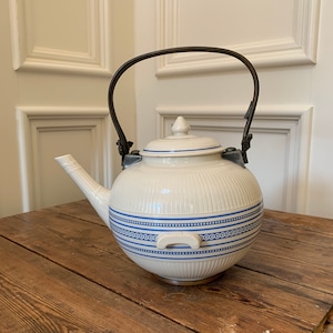 Puede incluir: Una tetera de cerámica blanca con un diseño de patrón azul y blanco y un asa de metal negro. La tetera está sentada en una mesa de madera.