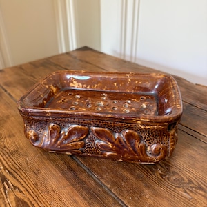 May include: A rectangular brown glazed ceramic soap dish featuring a raised leaf motif along the exterior sides and a series of drainage holes integrated into the base. The glossy finish has a mottled appearance with darker brown accents.