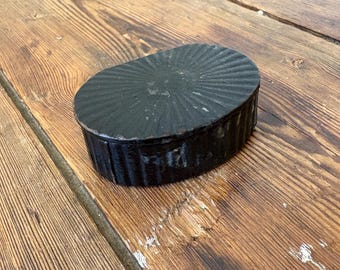May include: A small, black, oval-shaped box with a textured, ridged lid. The box sits on a weathered, wooden surface with visible grain and knots. The box's design features a series of vertical ridges around the sides and a sunburst pattern on the top.