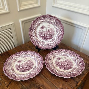 May include: Three antique decorative plates with a scalloped edge. The plates feature a detailed purple and white landscape design with buildings, trees, and figures. The plates are displayed on a wooden surface.