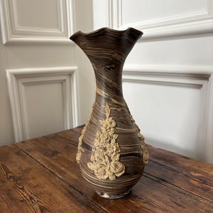 Pode incluir: Um vaso decorativo castanho e bege com uma parte superior com babados e detalhes de relevo floral. O vaso tem uma aparência marmoreada e fica sobre uma superfície de madeira. O vaso tem cerca de 30 cm de altura.
