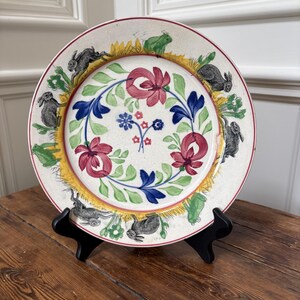 Puede incluir: Un plato de cerámica redondo con un diseño pintado a mano que presenta una corona central de flores rojas y azules con hojas verdes. El borde exterior muestra un patrón repetitivo de conejos grises y ranas verdes sobre un fondo de hierba amarilla.