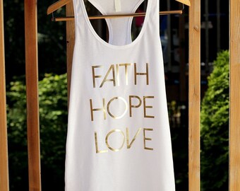 Puede incluir: Camiseta blanca de tirantes con las palabras "FAITH HOPE LOVE" impresas en dorado en la parte delantera.