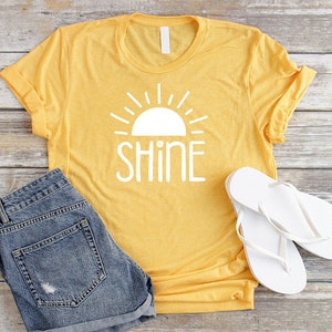 May include: A yellow t-shirt with a white sun and the word "shine" printed on the front. The shirt is paired with denim shorts and white flip flops.