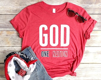 May include: Red t-shirt with white text that reads "GOD ONE NATION". The shirt is laid out on a wooden surface with a pair of sunglasses and a blue bandana.