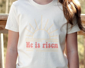 May include: A white t-shirt with a peach-colored sun and the text "He is risen" in a retro font.