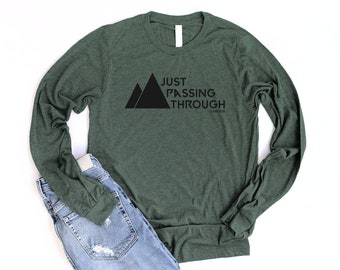 May include: A green long-sleeved shirt with the text "JUST PASSING THROUGH" and a graphic of two mountain peaks.