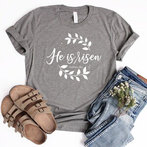 Puede incluir: Camiseta gris con el texto "He is risen" y un dise&ntilde;o de corona floral. El texto "Matthew 28:6" tambi&eacute;n est&aacute; incluido en la camiseta.