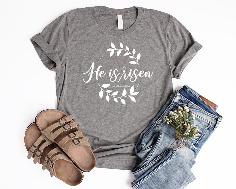 Puede incluir: Camiseta gris con el texto "He is risen" y un dise&ntilde;o de corona floral. El texto "Matthew 28:6" tambi&eacute;n est&aacute; incluido en la camiseta.