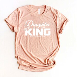 Op de afbeelding: Een perzikkleurig T-shirt met de tekst "Daughter of the King" en "2 Corinthians 6:18" in wit.