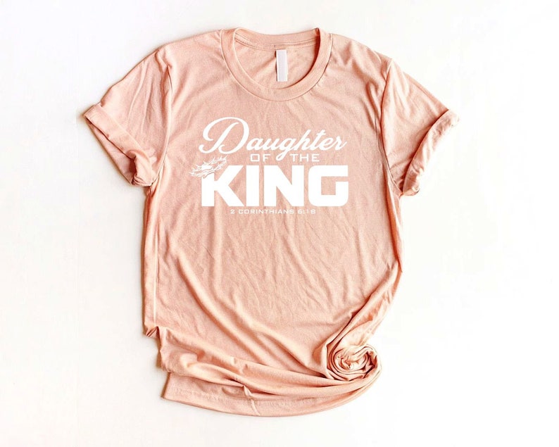 Op de afbeelding: Een perzikkleurig T-shirt met de tekst "Daughter of the King" en "2 Corinthians 6:18" in wit.