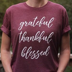 May include: A maroon t-shirt with rolled-up sleeves. The shirt features the words "grateful, thankful, blessed" in a white, cursive font. The shirt is made of a soft material and is suitable for casual wear.