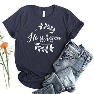 Puede incluir: Una camiseta azul marino con un dise&ntilde;o gr&aacute;fico blanco que presenta una corona de flores y el texto "He is risen" con la referencia b&iacute;blica "Mateo 28:6".