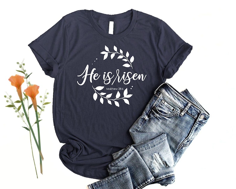 Puede incluir: Una camiseta azul marino con un dise&ntilde;o gr&aacute;fico blanco que presenta una corona de flores y el texto "He is risen" con la referencia b&iacute;blica "Mateo 28:6".