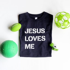 May include: A black t-shirt with the words "Jesus Loves Me" printed in white. The shirt is surrounded by green and white toys, including a football, a soccer ball, and a turtle.