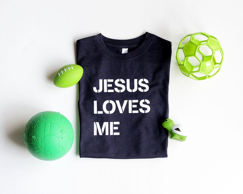May include: A black t-shirt with the words "Jesus Loves Me" printed in white. The shirt is surrounded by green and white toys, including a football, a soccer ball, and a turtle.