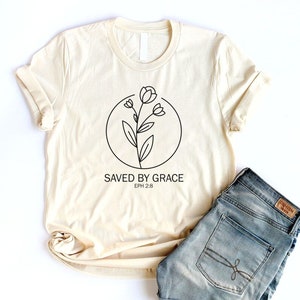 K&ouml;nnte beinhalten: Ein beiges T-Shirt mit einer schwarzen Linienzeichnung einer Blume in einem Kreis und dem Text "SAVED BY GRACE EPH 2:8".