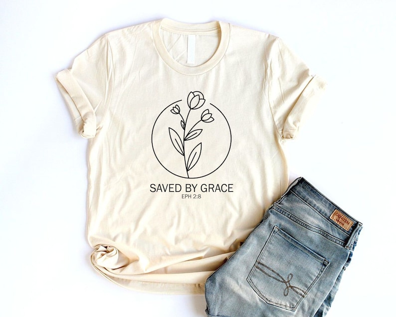 K&ouml;nnte beinhalten: Ein beiges T-Shirt mit einer schwarzen Linienzeichnung einer Blume in einem Kreis und dem Text "SAVED BY GRACE EPH 2:8".