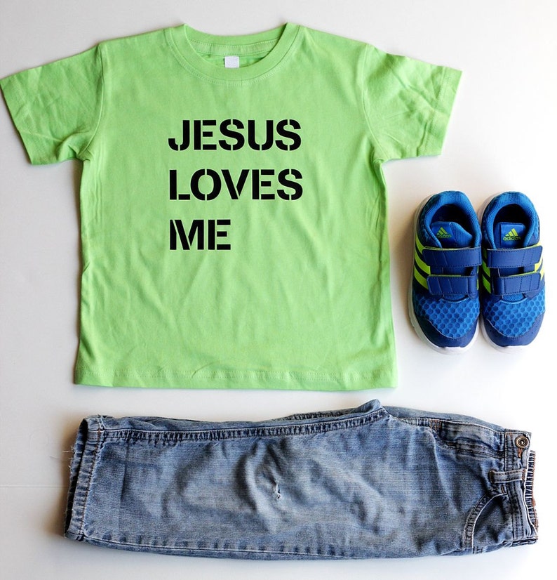May include: A lime green t-shirt with the text "JESUS LOVES ME" in black lettering. The shirt is paired with blue jeans and blue and green sneakers.