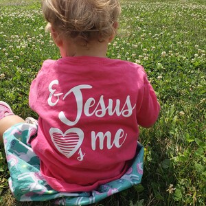 May include: A young child wearing a pink t-shirt with the words "& Jesus loves me" and a white heart graphic. The child is sitting in a field of clover.
