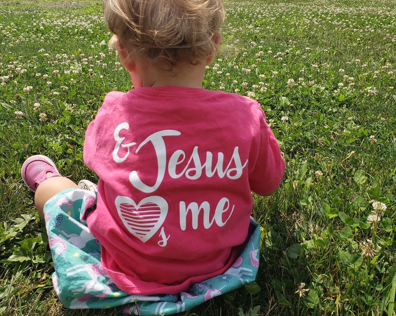 May include: A young child wearing a pink t-shirt with the words "& Jesus loves me" and a white heart graphic. The child is sitting in a field of clover.