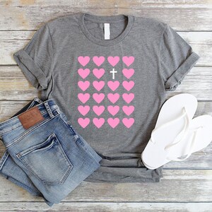 May include: A gray t-shirt with a pink heart design and a white cross in the center. The hearts are arranged in a grid pattern.