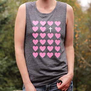 K&ouml;nnte beinhalten: Ein graues &auml;rmelloses Shirt mit einem rosafarbenen Herzdesign und einem wei&szlig;en Kreuz in der Mitte. Das Shirt hat eine lockere Passform und einen runden Ausschnitt.