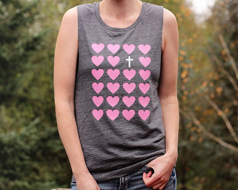 K&ouml;nnte beinhalten: Ein graues &auml;rmelloses Shirt mit einem rosafarbenen Herzdesign und einem wei&szlig;en Kreuz in der Mitte. Das Shirt hat eine lockere Passform und einen runden Ausschnitt.